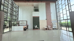Um hall amplo com balcão de recepção, parede verde decorativa e grandes janelas com vista para o exterior.