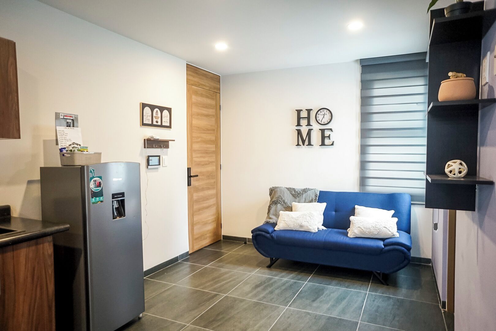 Una cocina moderna con un refrigerador, una puerta de madera, un sofá azul con cojines y una estantería con adornos.