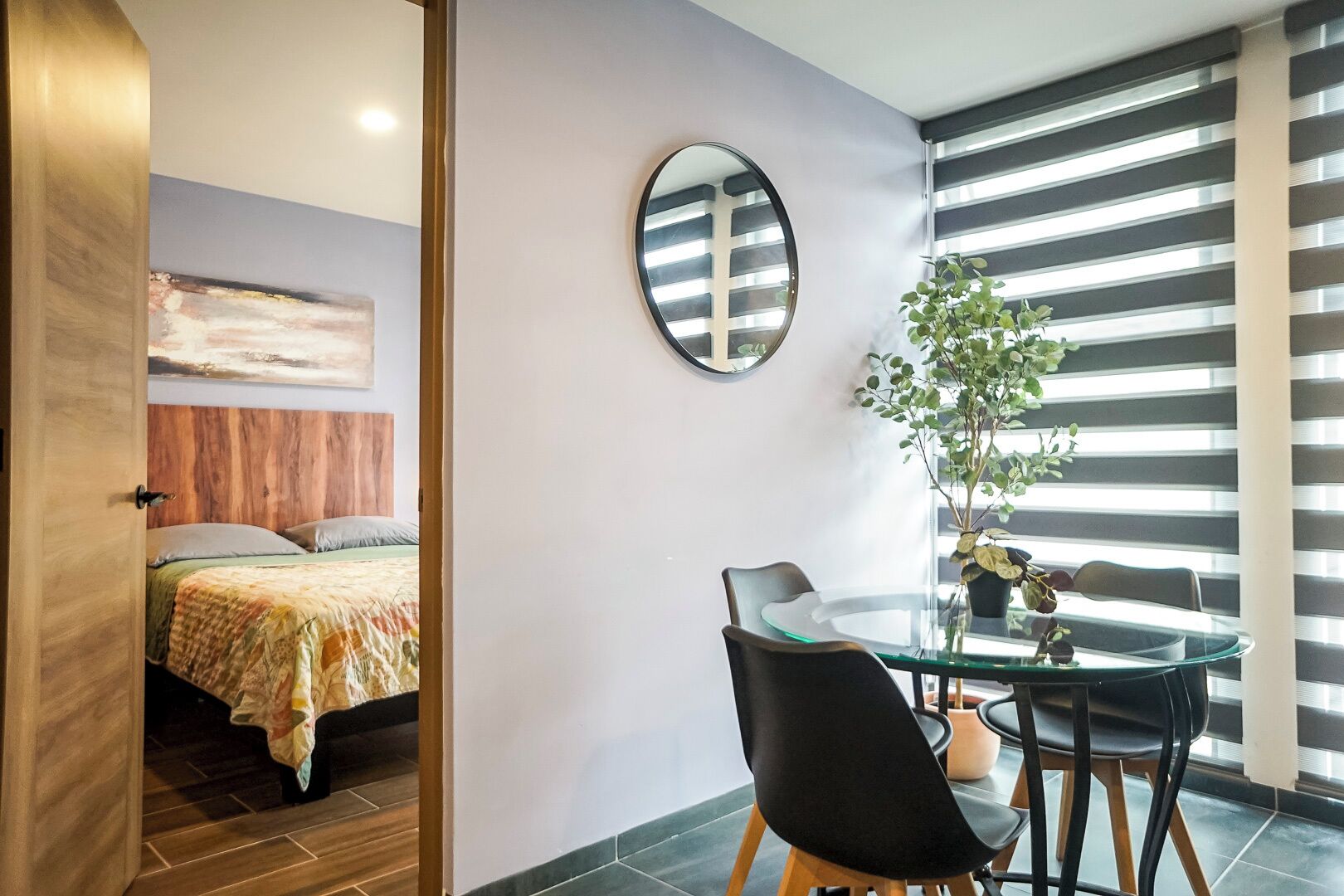 Una habitación de hotel moderna con cama, zona de comedor con mesa y sillas de vidrio, un espejo grande y una planta en maceta.