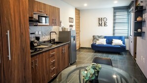 A modern kitchen with wooden cabinets, a refrigerator, and a coffee maker. There is a blue sofa in the living area with a 'HOME' sign on the wall. - 1BR Flat with Smart Access & Parking (San Luis Potosi)