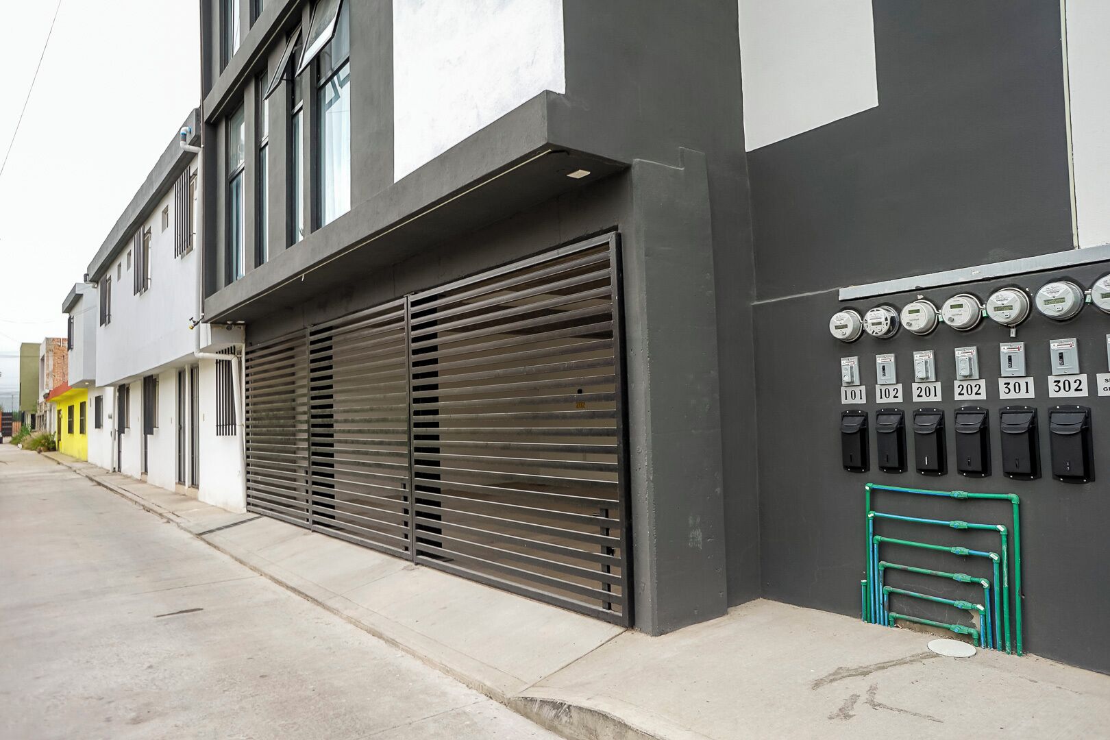 Un bâtiment moderne doté d’un grand portail de garage, d’une rangée de boîtiers électriques numérotés et d’un tuyau vert posé sur le sol.