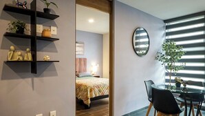 A modern hotel room with a bed, a dining area with a table and chairs, a mirror, and shelves with decorative items. - 1BR Flat with Smart Access & Parking (San Luis Potosi)