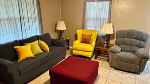 A living room with a dark sofa, a red ottoman, a yellow armchair, and a grey armchair. There are two lamps on side tables. - Charming 3-bedroom, spacious home conveniently located near Tuskegee University (Tuskegee)