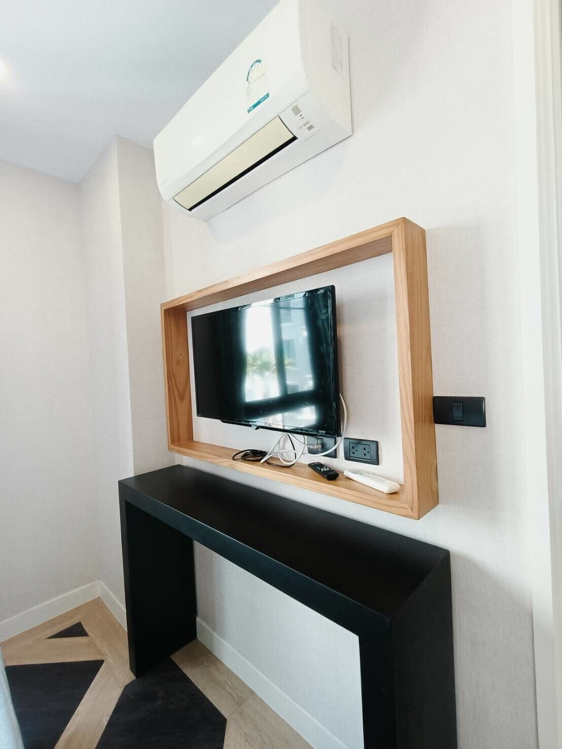 A modern living room with a flat-screen TV mounted on a wooden frame, a black entertainment unit below, and an air conditioning unit above.
