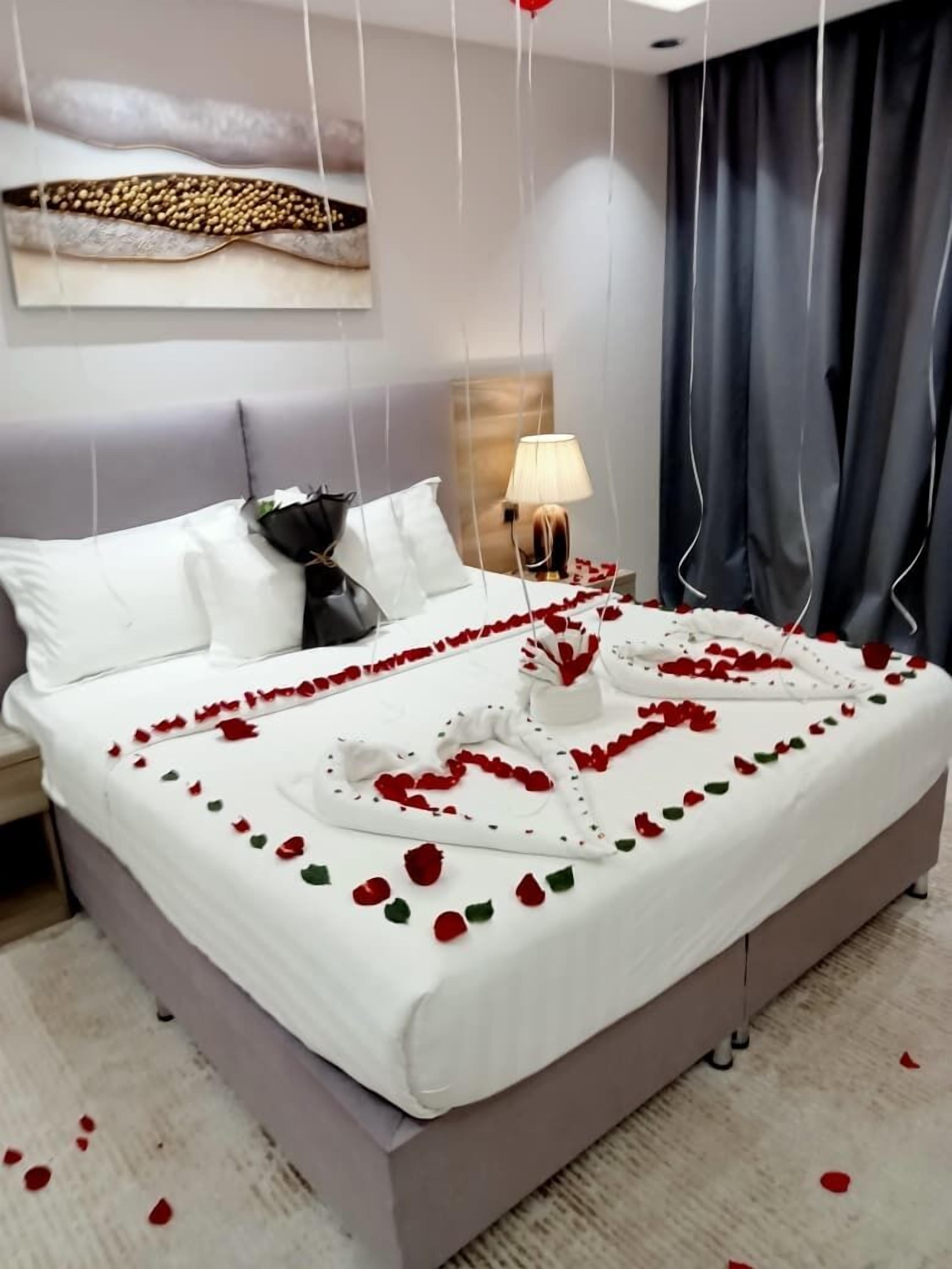 A neatly made bed with rose petals and a heart-shaped decoration, a lamp on the nightstand, and a modern headboard.