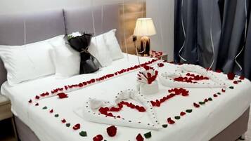 A neatly made bed with rose petals and a heart-shaped decoration, a lamp on the nightstand, and a modern headboard.