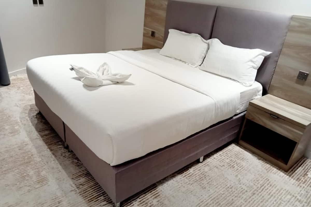 Ein ordentlich bezogenes Bett mit weißer Decke und Kissen, ein hölzernes Kopfteil und ein kleiner Nachttisch mit Schublade.