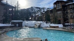 Une piscine avec une personne à l'intérieur, entourée de bâtiments recouverts de neige et avec une chaîne de montagnes en arrière-plan.