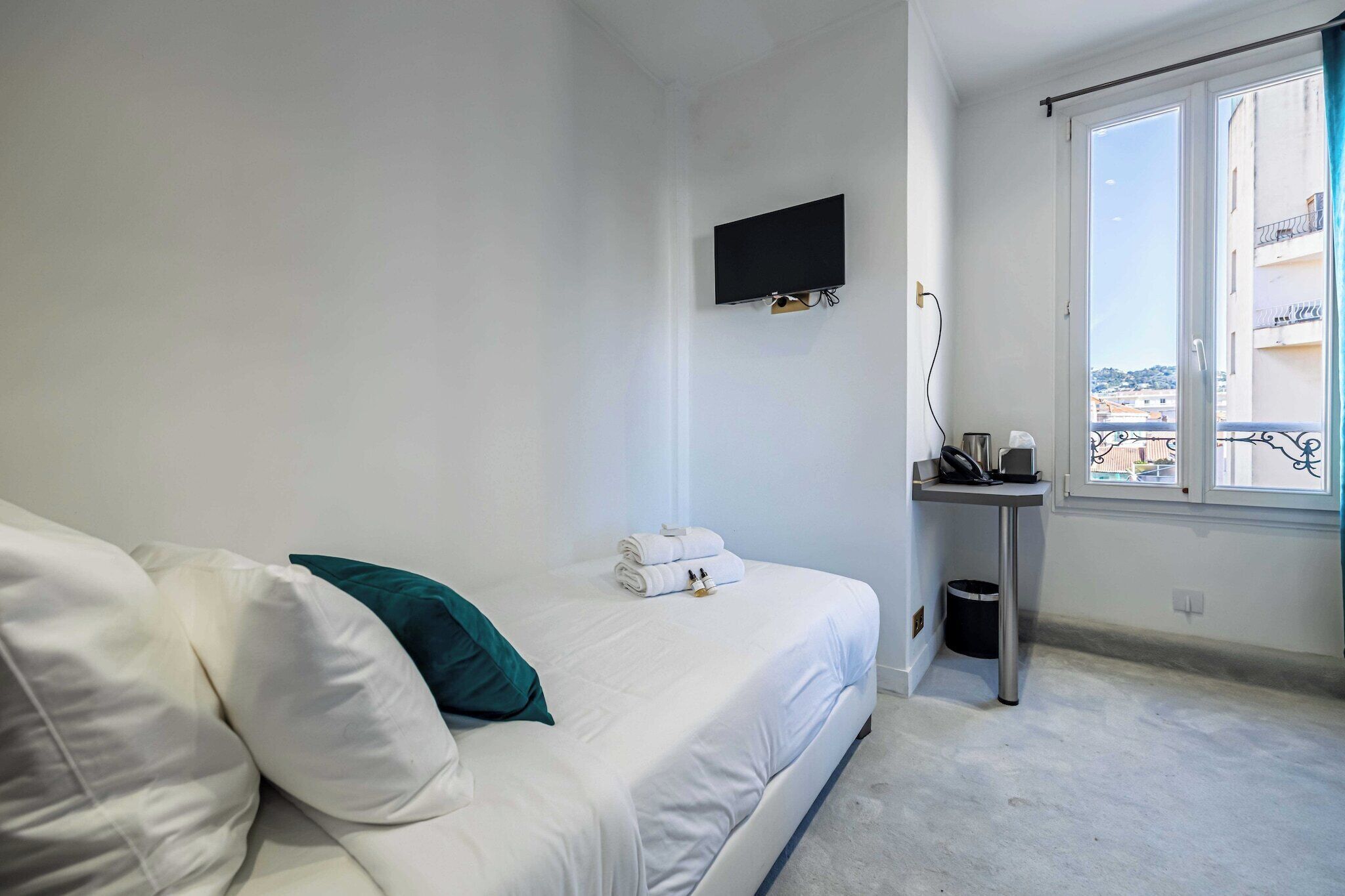 Ein Schlafzimmer mit Bett, einem an der Wand montierten Fernseher, einem kleinen Tisch mit Mülleimer und einem Fenster mit Blick auf Gebäude.