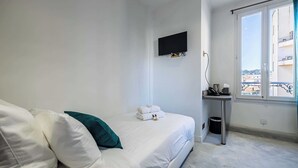 Ein Schlafzimmer mit Bett, einem an der Wand montierten Fernseher, einem kleinen Tisch mit Mülleimer und einem Fenster mit Blick auf Gebäude.