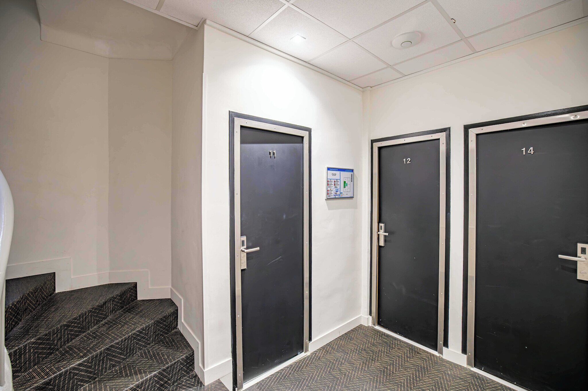 A hallway with three doors, numbered 12, 13, and 14, leading to rooms. There is a small sign on the wall between doors 12 and 13.
