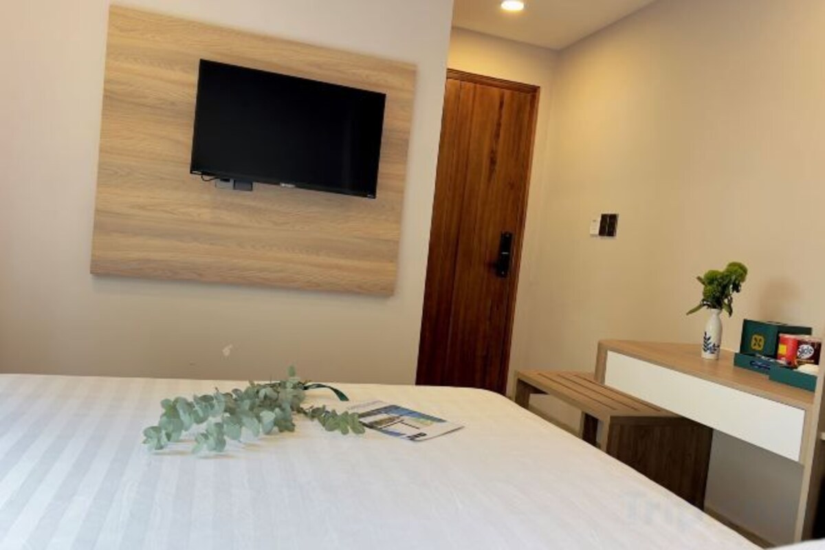 A hotel room with a bed, a mounted TV, a wooden door, and a small desk with a vase and a box.