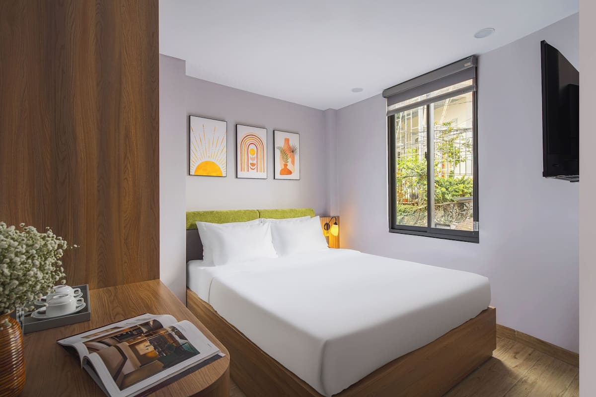 Moderni hotellihuone, jossa on suuri sänky, puinen pääty, ikkuna, josta avautuu näkymä vehreään maisemaan, sekä kolme kehystettyä taideteosta seinällä.