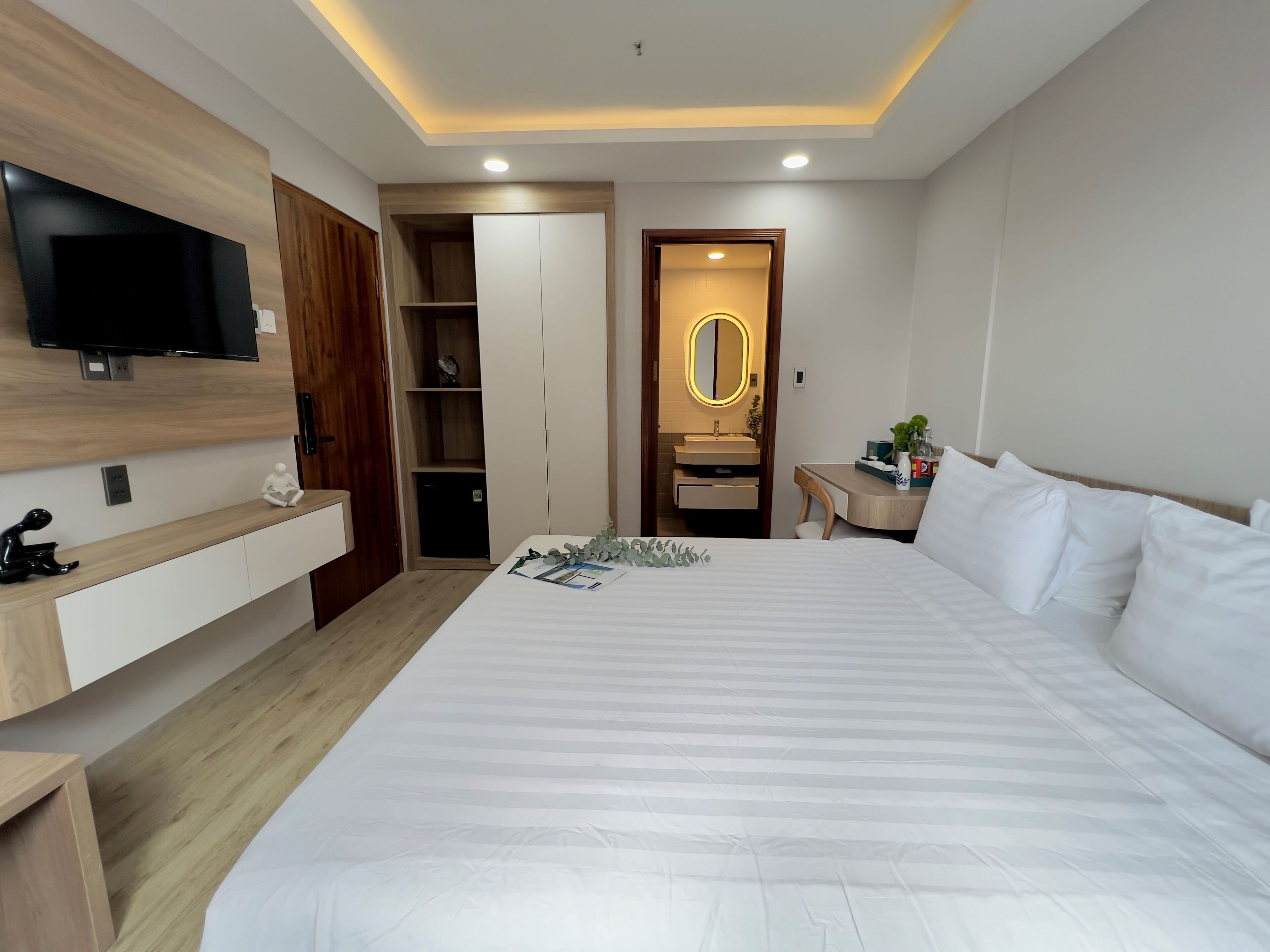 Une chambre d’hôtel moderne dotée d’un grand lit, d’un téléviseur fixé au mur, d’une armoire en bois, d’une armoire intégrée, d’une coiffeuse avec un miroir et d’une petite table avec des articles de toilette.