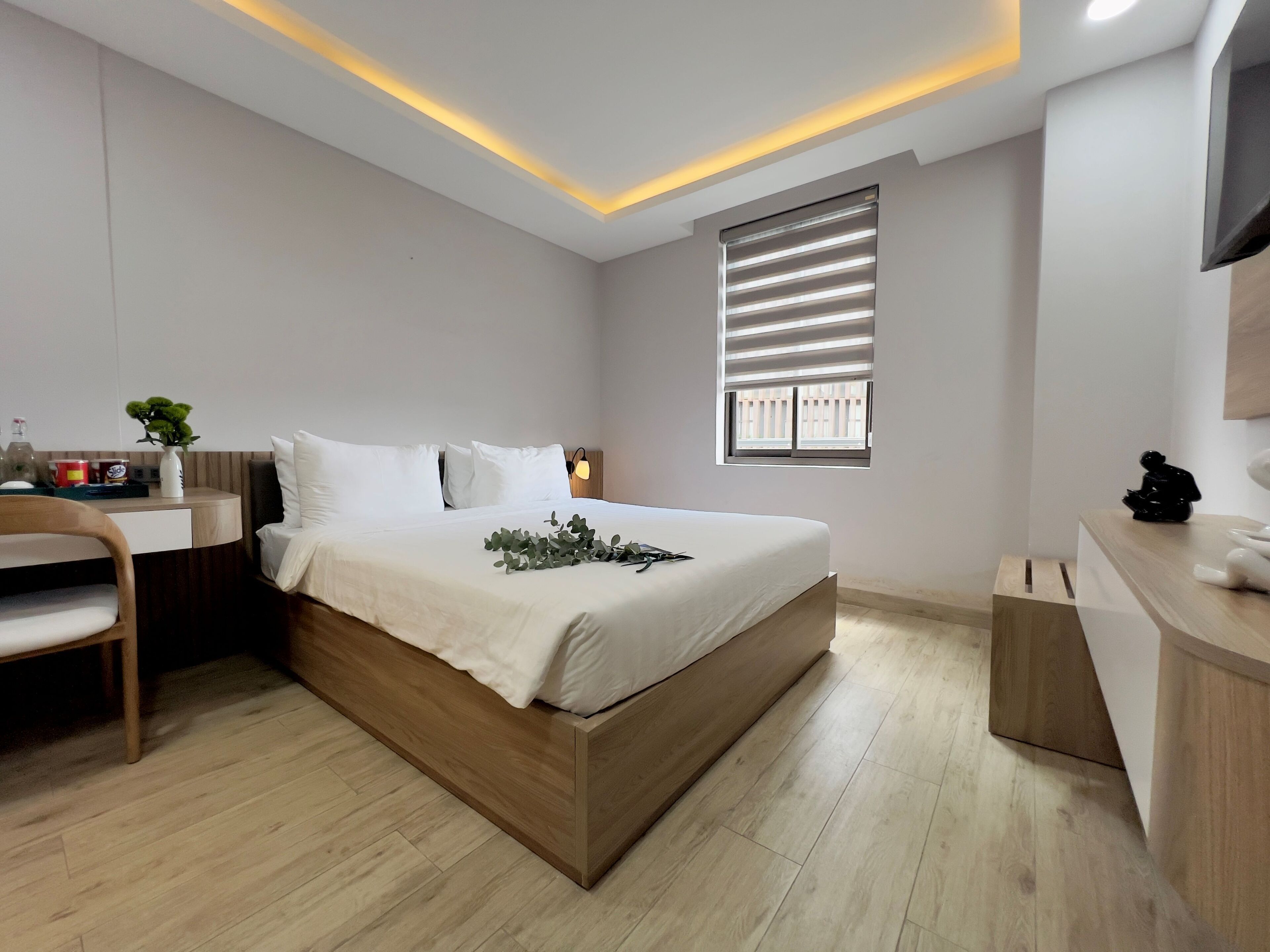 A modern bedroom with a wooden bed frame, a desk with a chair, a bedside table, and a window with blinds.