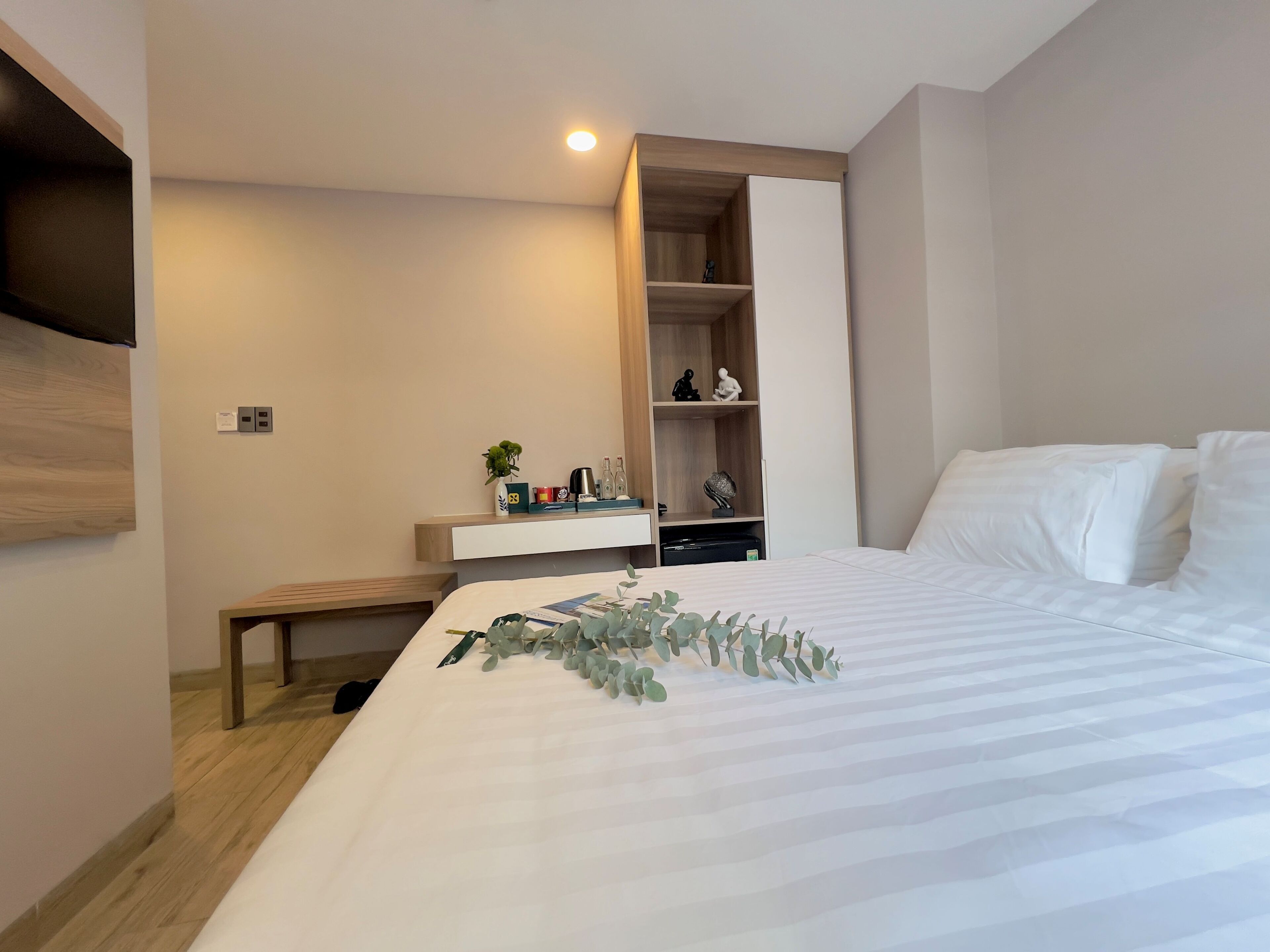 Een moderne slaapkamer met een groot bed, een nachtkastje, een bankje, een garderobe en een klein plankje met decoratieve objecten.