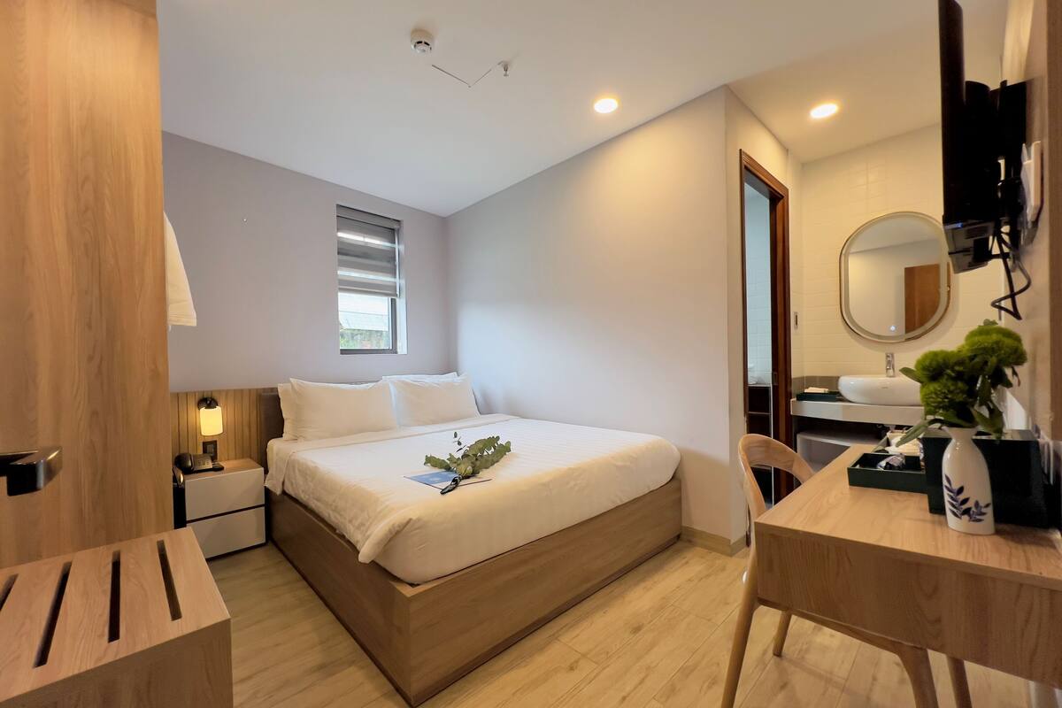 Moderni hotellihuone, jossa on puinen sänky, yöpöytä ja työpöytä tuolilla. Huoneessa on ikkuna, jossa on sälekaihtimet, peili ja pieni kasvi työpöydällä.