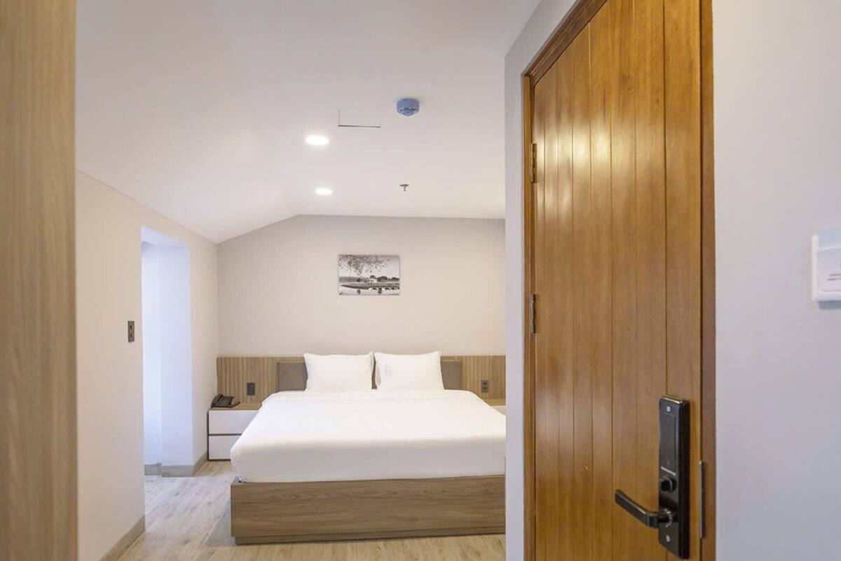 Hotellihuone, jossa on puinen ovi, valkoisilla lakanoiden peitetty sänky, yöpöytä ja seinälle ripustettu taulu.