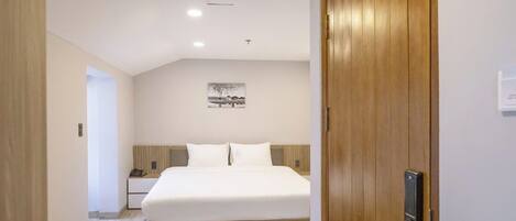 Een hotelkamer met een houten deur, een bed met wit beddengoed, een nachtkastje en een aan de muur hangend schilderij.