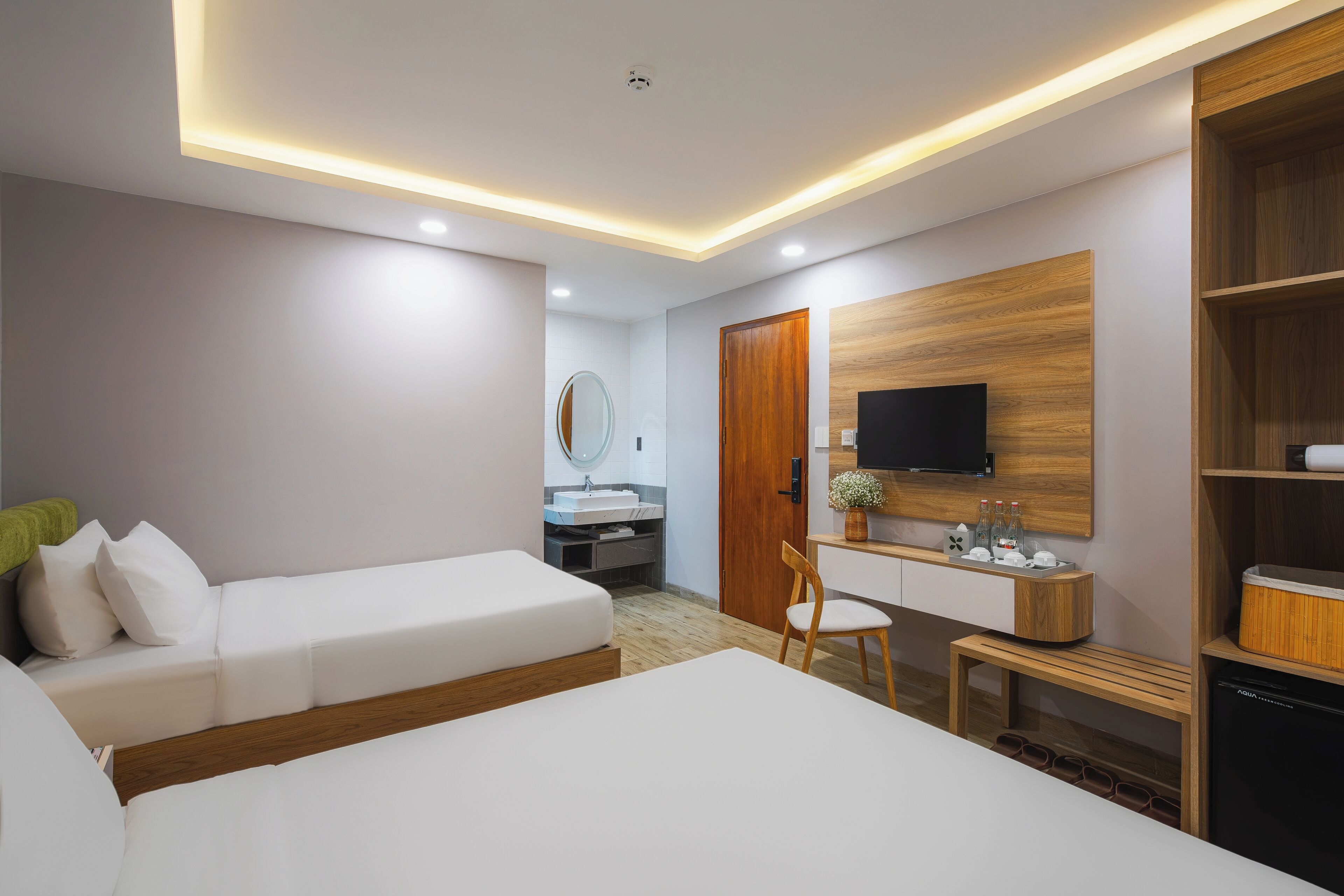 A modern hotel room with two beds, a desk, a mirror, a TV, and a wooden bookshelf.