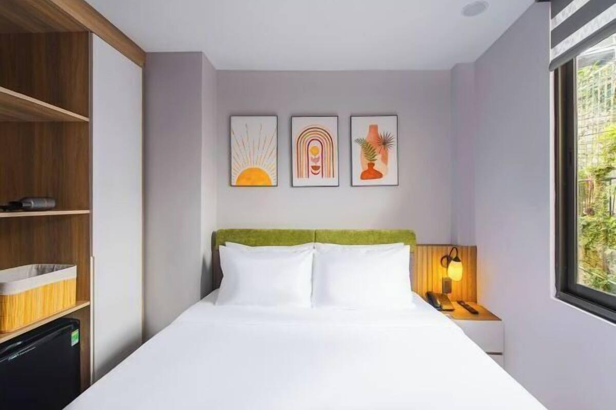 Hotellihuone, jossa on sänky, yöpöytä, lamppu ja kolme kehystettyä taideteosta seinällä.