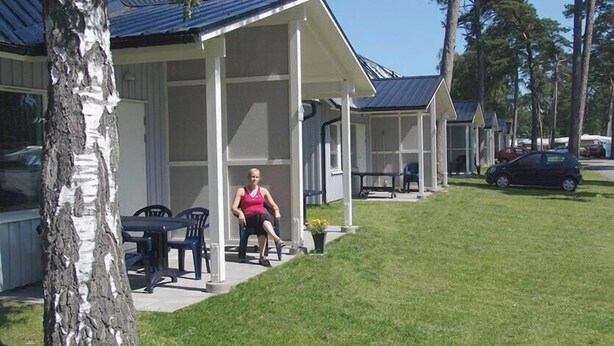 A row of single-story cabins with a covered porch, a grassy lawn, and a car parked nearby. - Ystad Camping (Ystad)