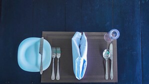 Ein Tisch gedeckt mit einem blauen Teller, Besteck, einer Serviette, einem Glas und einer Flasche auf dunklem Holztisch.
