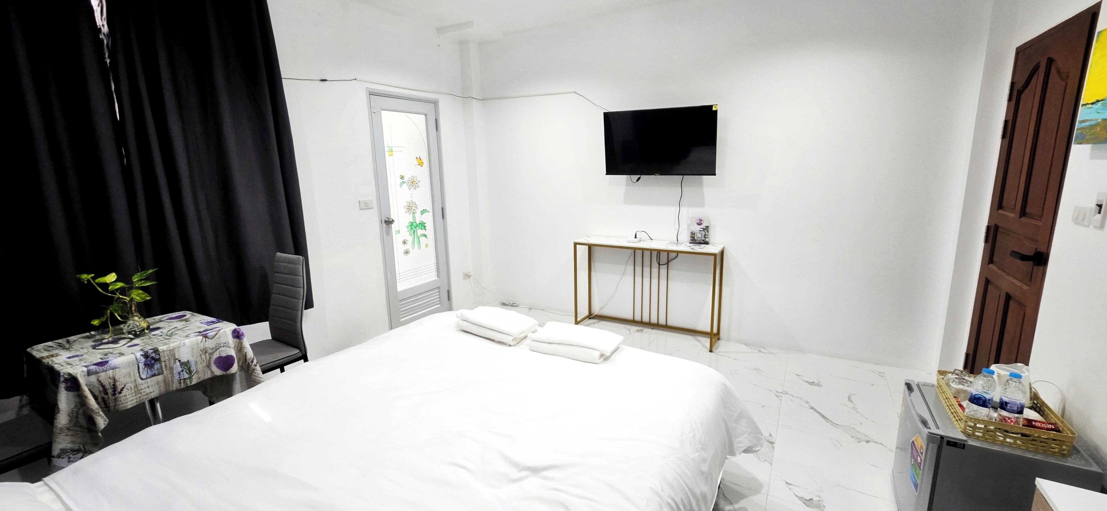 Une chambre d’hôtel avec un lit, une petite table sur laquelle est posé un vase, un téléviseur fixé au mur et une porte dotée d’une vitre en verre dépoli.