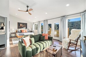 A spacious living room with a green sectional sofa, a wooden rocking chair, and a large window with curtains.