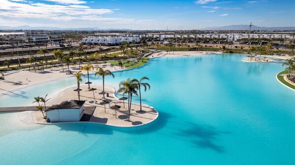 Una piscina grande y cristalina de color azul con una zona de playa arenosa, palmeras y una pequeña isla con una estructura de techo de paja.