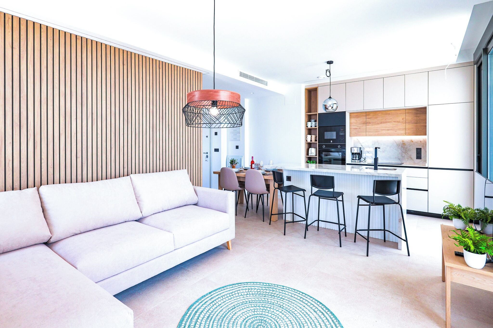 A modern living room with a white sofa, a dining area with chairs, a kitchen with white cabinets, and a wooden wall.