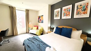 A hotel room with a bed, a desk, a chair, a window with curtains, and three framed posters on the wall.