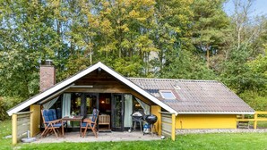Una cabaña pequeña de un solo nivel con techo inclinado, chimenea y un patio con mobiliario exterior.