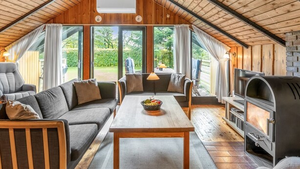 Una habitación acogedora con paneles de madera, techo alto, chimenea, sofá, mesa de comedor y ventanas amplias.