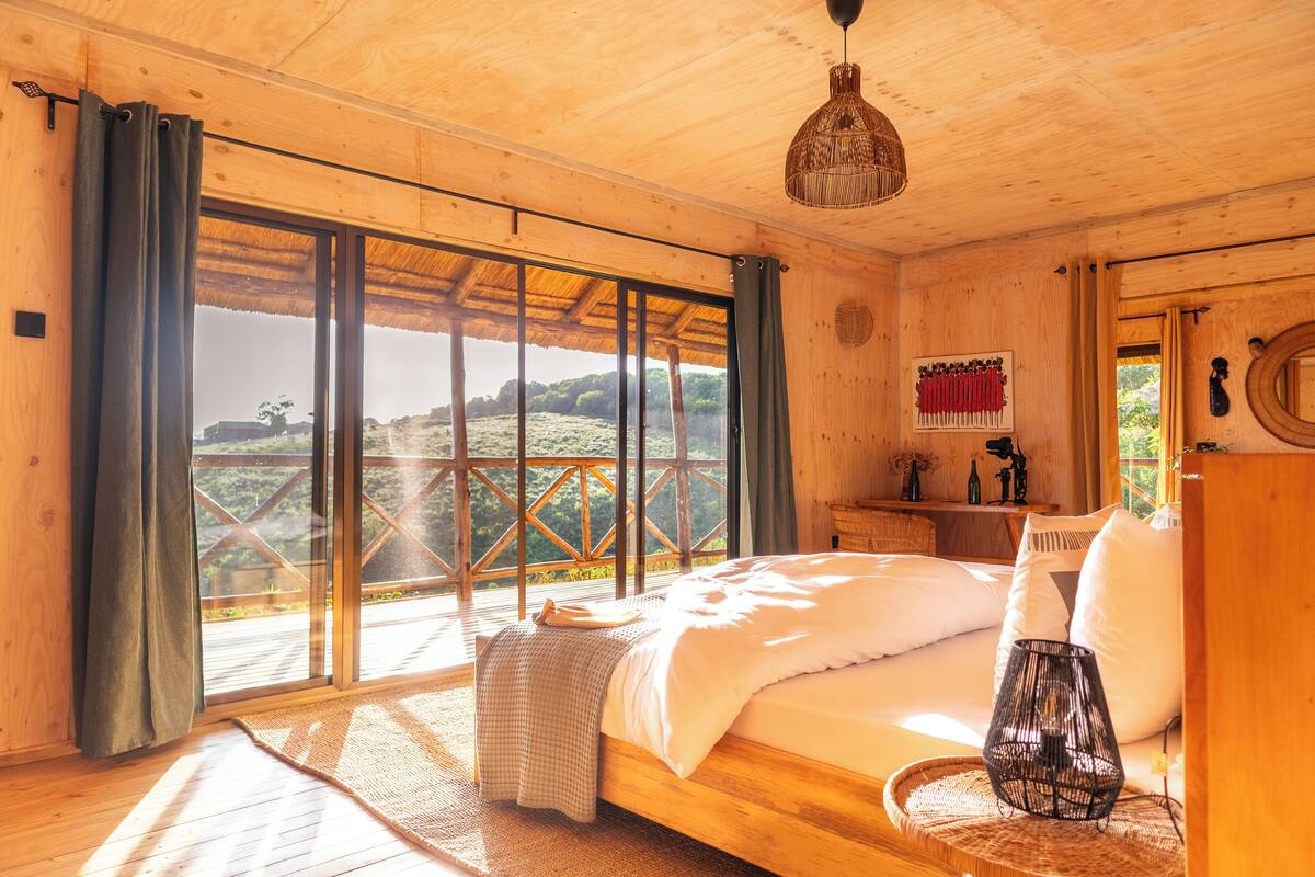A wooden bedroom with a large bed, a nightstand, a lamp, and a view of the outdoors.