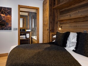 A bedroom with a wooden headboard, a bed with a dark blanket, a nightstand with a lamp, and a bathroom visible through an open door.