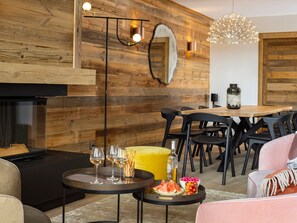 A modern living room with a wooden wall, a fireplace, a dining area with a table and chairs, and a cozy seating area with a sofa and ottoman.