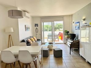A modern kitchen with a dining area, a sofa with colorful cushions, a floor lamp, and a large window with a view of greenery.