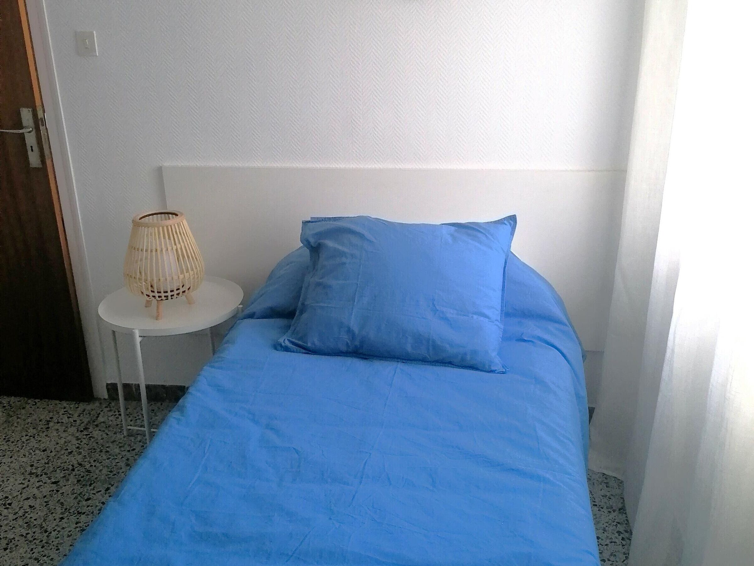 A single bed with a blue mattress and pillow, a small round table with a lamp, and a white wall.