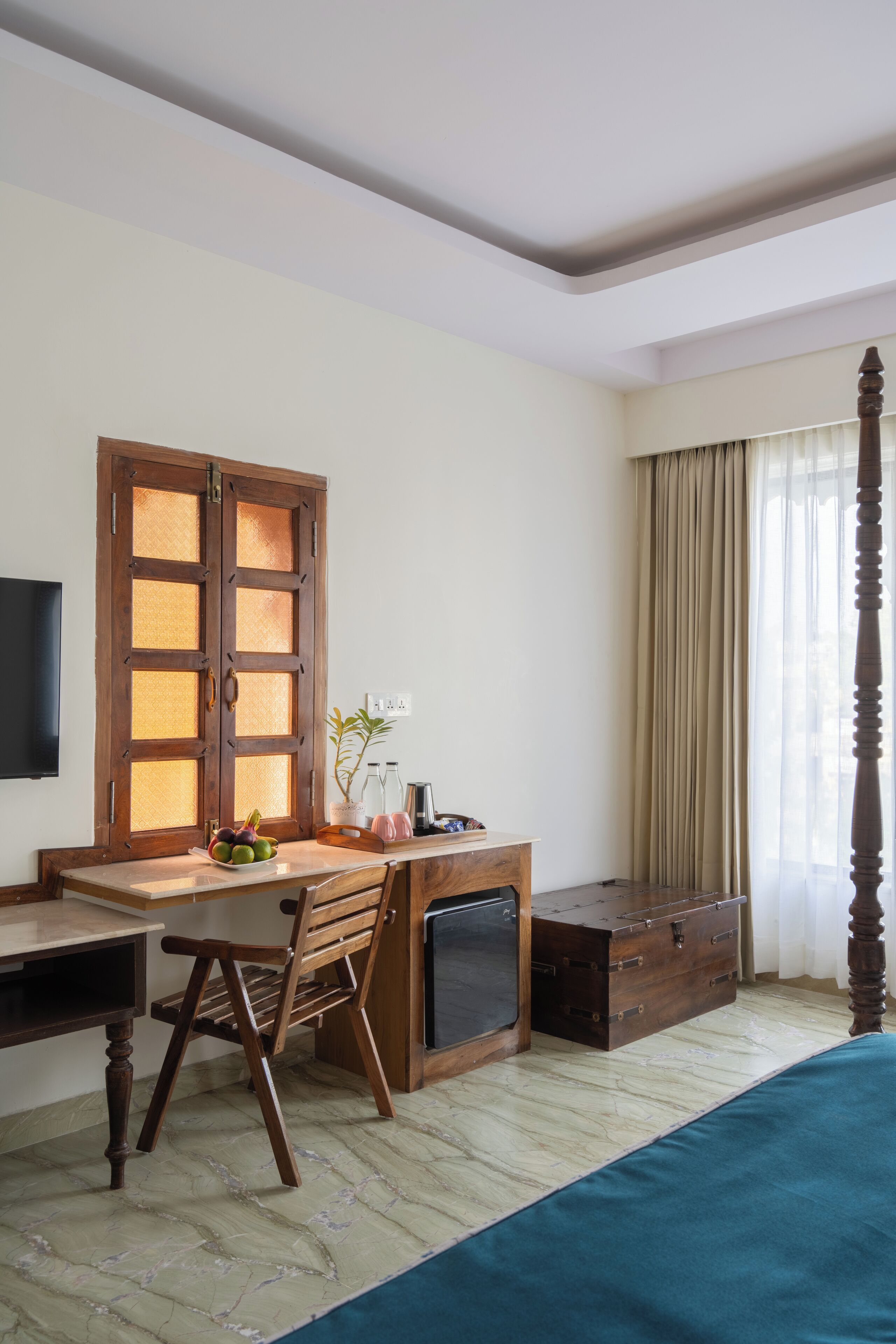 Una habitación con un escritorio de madera, una silla, un mueble pequeño, un televisor montado en la pared y un ventanal con cortinas.