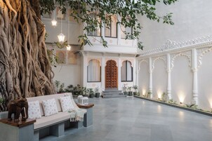 Un amplio patio con un tronco de árbol grande, un banco de madera y un edificio tradicional con puertas y columnas ornamentadas.