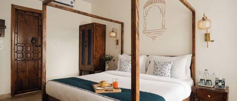 Una cama con dosel de madera, ropa de cama blanca y un plaid color turquesa. Hay una mesita de noche de madera con un decantador de vidrio y una pequeña bandeja con un vaso de jugo de naranja. Se observa un armario de madera al fondo.