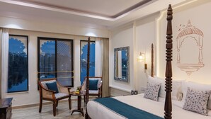 A four-poster bed with a canopy, a sitting area with chairs, a small table with fruit, and a large window with a view of the cityscape. - The Experience Resort (Udaipur)