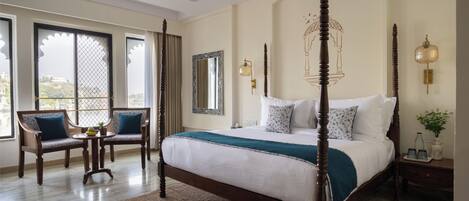A four-poster bed with a white and teal bedspread, two upholstered chairs with blue cushions, a small round table with a fruit bowl, and a large window with a view of buildings.