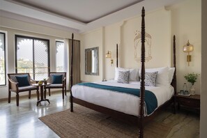A four-poster bed with a white and teal bedspread, two upholstered chairs with blue cushions, a small round table with a fruit bowl, and a large window with a view of buildings. - The Experience Resort (Udaipur)