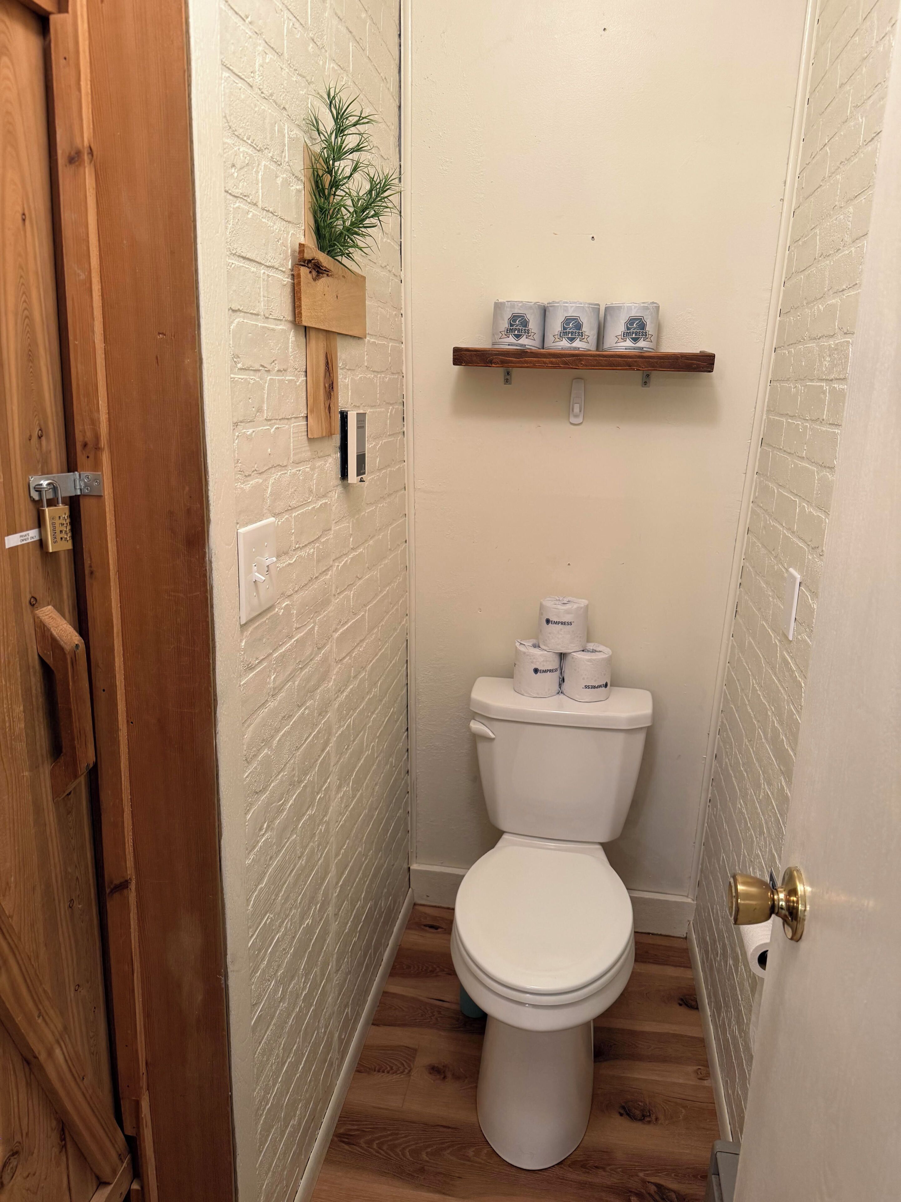 A small, narrow bathroom with a white toilet, a wooden shelf with toilet paper, and a door with a brass handle.