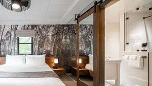 Quarto de hotel moderno com uma cama grande, uma mesinha de cabeceira de madeira, uma mesa de apoio com abajur, um espelho e um banheiro com chuveiro e porta-toalhas.