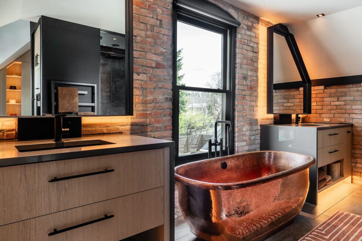 A modern bathroom with a large copper sink, a black-framed mirror, and a view of the outdoors through a large window.