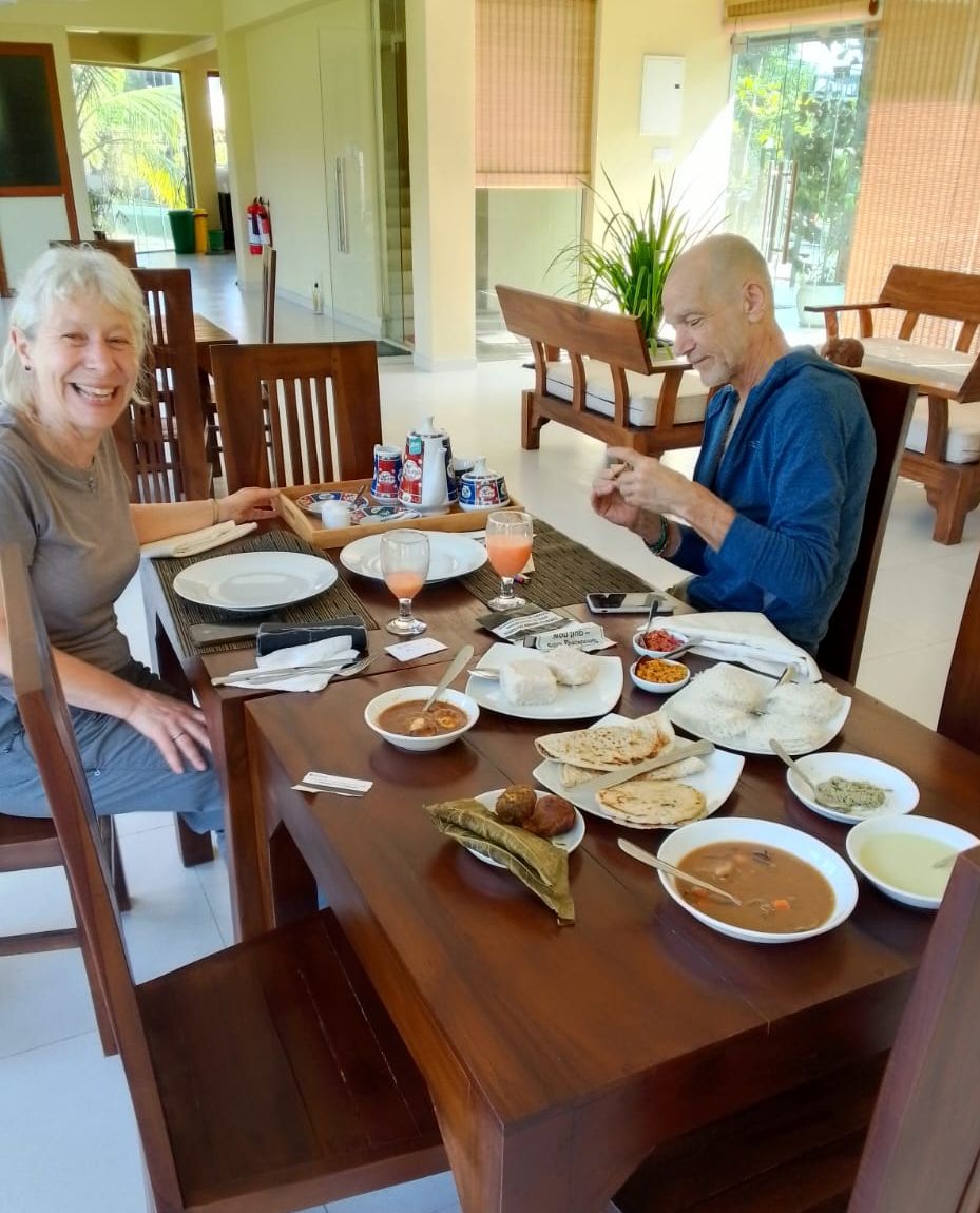 A dining table with various dishes, including rice, soup, and flatbreads, in a room with wooden furniture and a plant in the background.