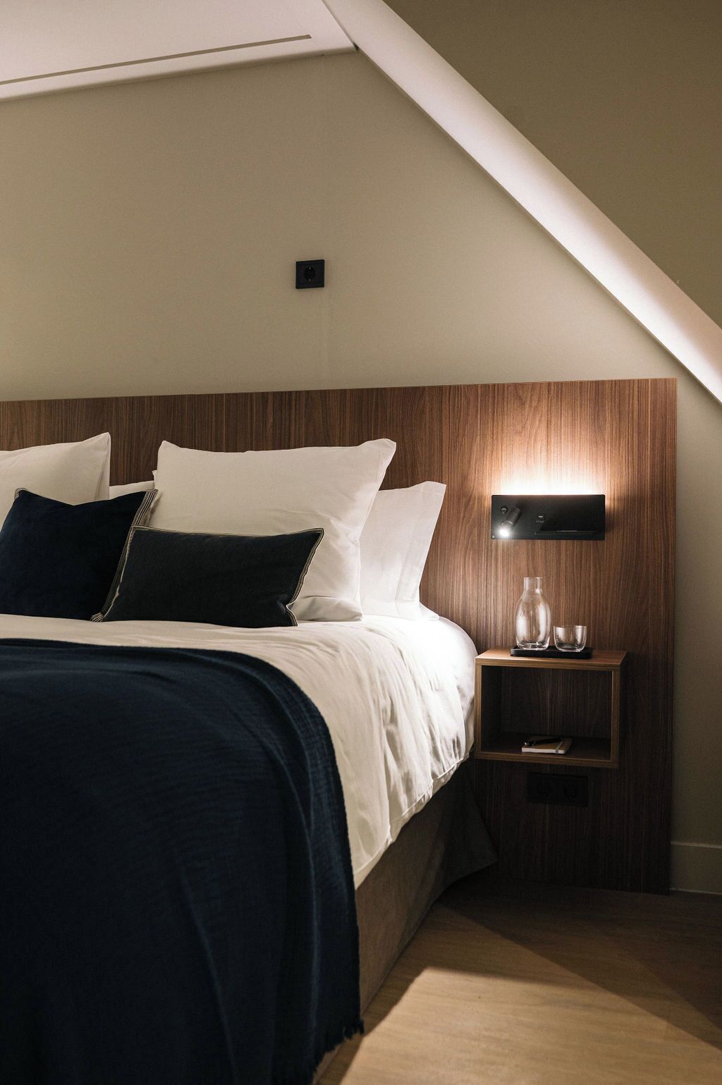 A modern bedroom with a sloped ceiling, a wooden headboard, a bedside table with a lamp, and a nightstand.